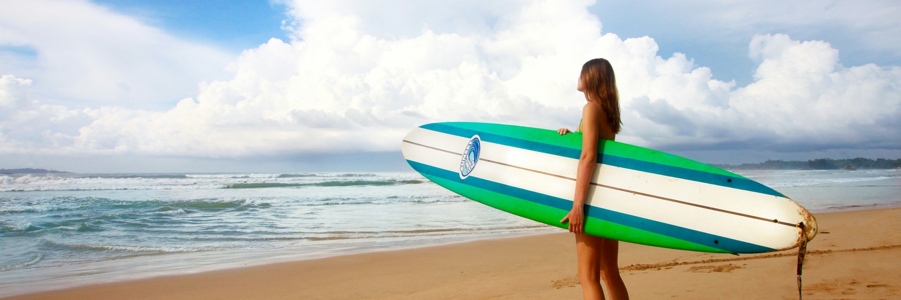 Woman holding a surfboard on a sandy beach in Weligama, enjoying surf in Sri Lanka with ocean waves and cloudy sky in the background